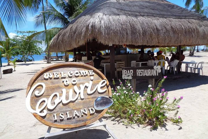 Schild „Willkommen auf Cowrie Island“ mit einer Strohdach-Bar im Hintergrund